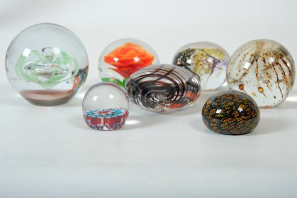 SIEBEN PAPERWEIGHTS, von klein bis groß: kleines durchsichtiges Paperweight mit Blumen im inneren, 115g. Kleines schwarzes Paperweight mit Blumen aufgemalt, 139g. Mittleres durchsichtiges Paperweight mit einer schwarzen, einer violetten und einer gelben Blume, 439g. Mittleres durchsichtiges Paperweight mit einer Rose, 414g. Mittleres Paperweight mit Lufteinschlüssen, braun, 444g. Herzförmiges Paperweight mit violetten Streifen, 370g. Größeres Paperweight mit lilienähnlichem Lufteinschluss, gr