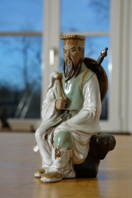 Kriegerfigur, chinesisch. Keramik. Sitzender Krieger, Schwert und Schild auf dem Rücken tragend, in der Hand einen Fächer. 18 x 8 x 9 cm.