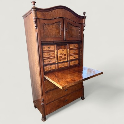 Louis Philippe Schreibschrank, um 1860. Fünfschübiger Sekretär mit quer-oval kassettierter Schreibklappe und Schrankelement, auf Klotzfüßen. Mittelfach mit jagdlichen Intarsien verziert, flankiert von jeweils vier seitlichen Schüben und einem Sockelschub, verziert mit ornamentalen Intarsieren. Die Rückwand des Mittelfachs durch 